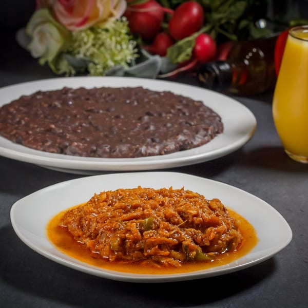 Two plates of Caribbean food: a dark brown bean dish and a vibrant orange stew, with a drink.