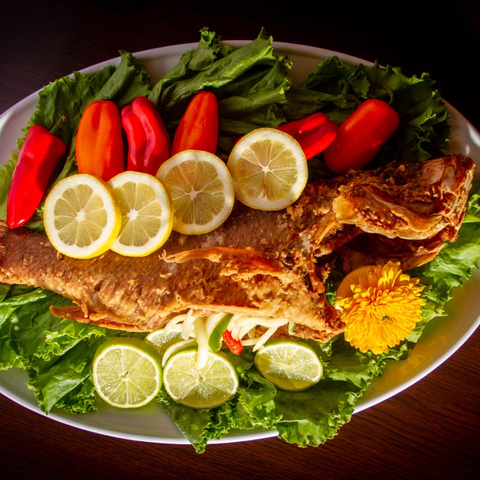 A whole fried fish, garnished with lemons, limes, red peppers, and a yellow flower on lettuce.